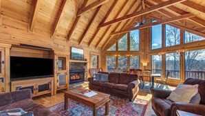 A spacious living room with a fireplace, wooden ceiling beams, and large windows offering a view of the outdoors. - Spacious 4BD Cabin w/ Hot Tub! Fireplaces + GAMES! (Sevierville)
