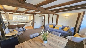A cozy living area with a wooden table, a sofa with yellow cushions, and a dining area with chairs. - Dove Cott | Sleeps 4 | Central Location (Croyde)