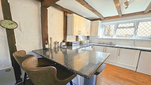 A kitchen with a central island, wooden beams, white cabinets, and a window with a view. - Dove Cott | Sleeps 4 | Central Location (Croyde)