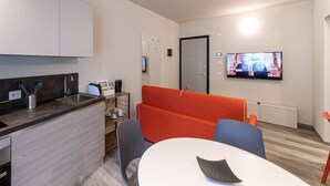Quarto de hotel moderno com cozinha compacta, área de estar com sofá e poltronas, televisão de tela plana na parede e porta com puxador.