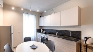 Cozinha moderna com armários brancos, bancada preta e forno embutido. Há uma mesa de jantar com cadeiras e uma janela com cortinas.