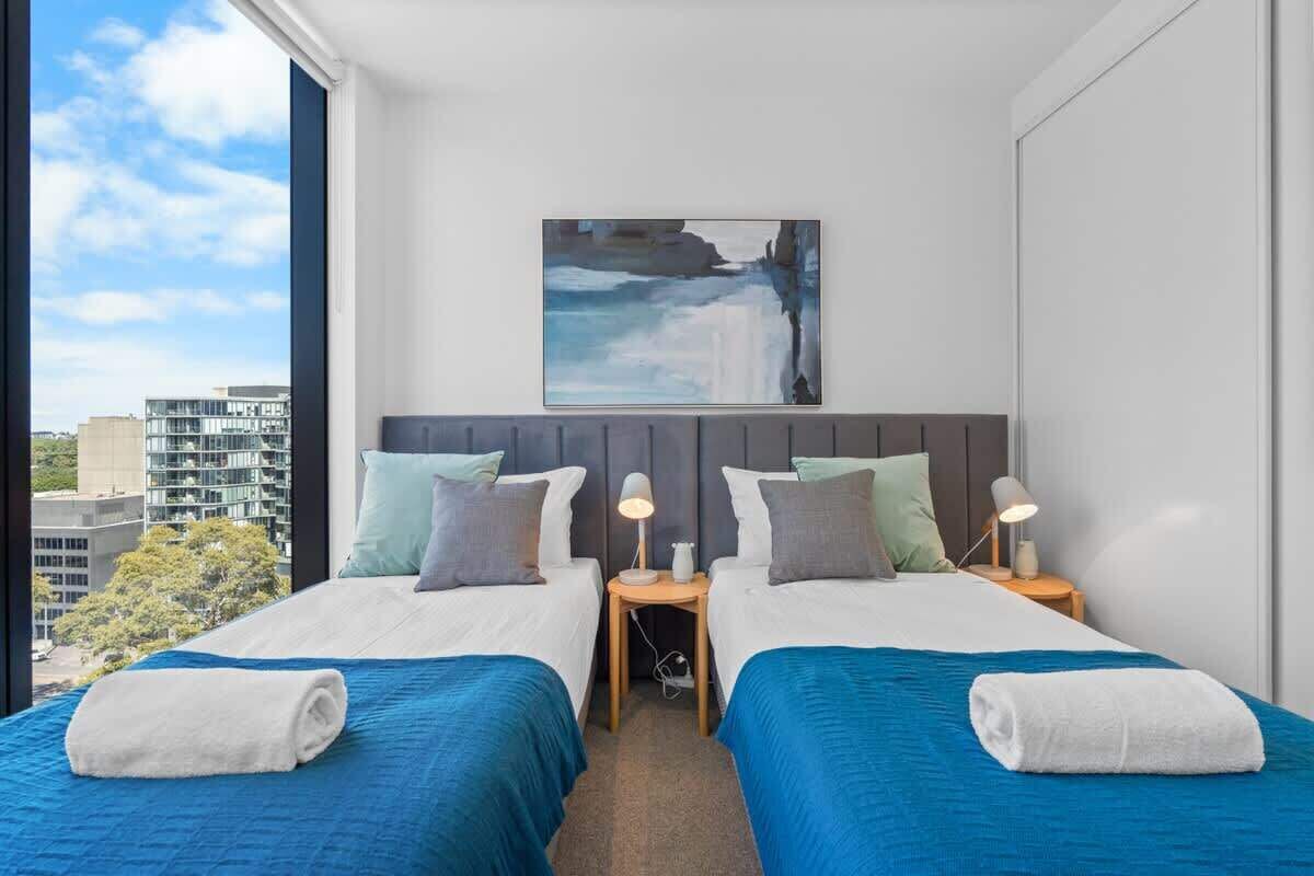 Een hotelkamer met twee bedden, uitzicht over de stad, een schilderij aan de muur en een raam met uitzicht op gebouwen en bomen.