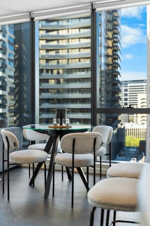 A modern dining area with a glass table and four chairs, large windows with a city view, and a balcony with a railing. - Brand New 2BR Oasis Retreat w/ Parking, Pool & Gym (South Melbourne)