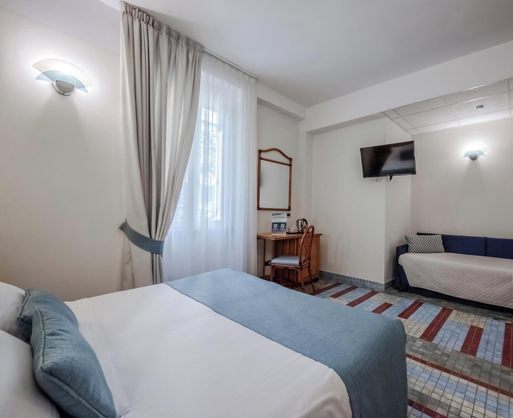 A hotel room with a bed, a desk with a chair, a television, and a window with curtains.