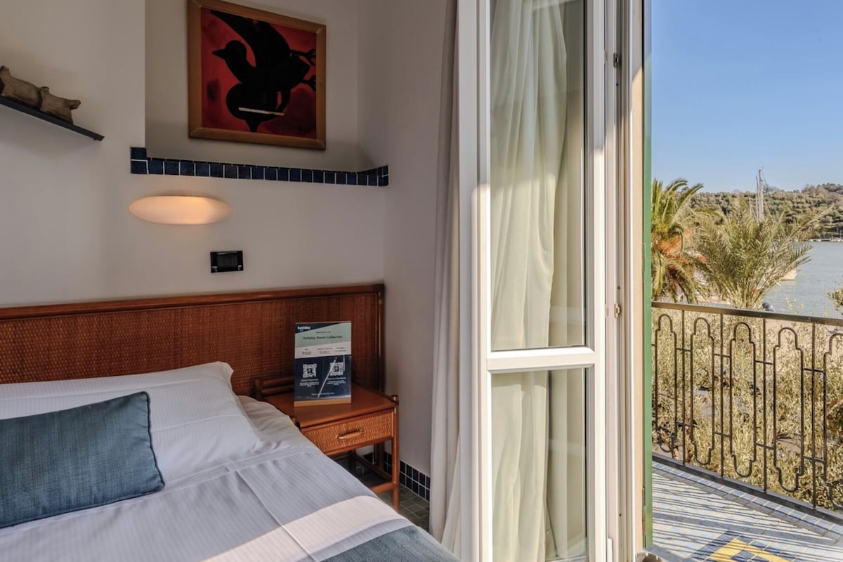 Habitación de hotel con cama, mesita de noche, una obra de arte en la pared, ventana con vista a un cuerpo de agua y palmeras, y balcón con barandilla.