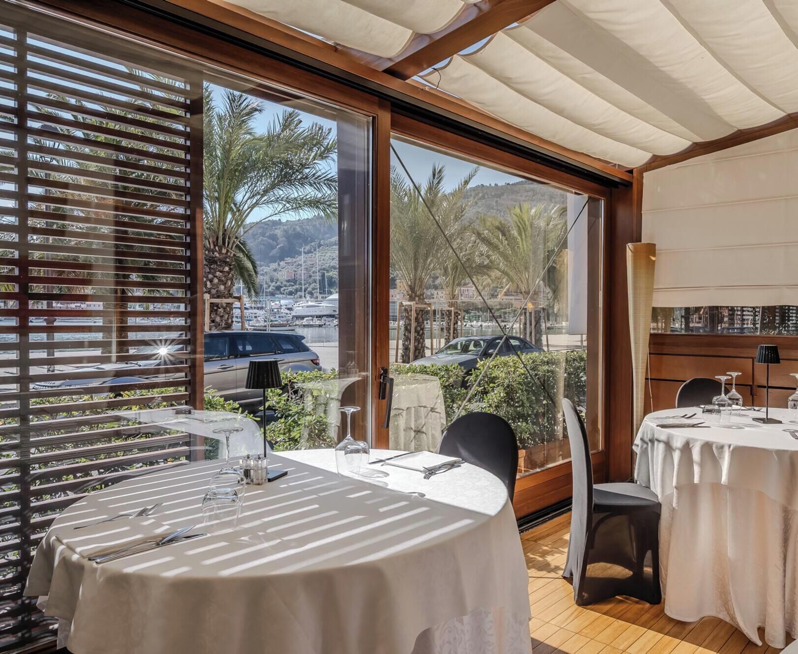 A dining area with tables set for two, large windows with a view of a marina, and a wooden ceiling.