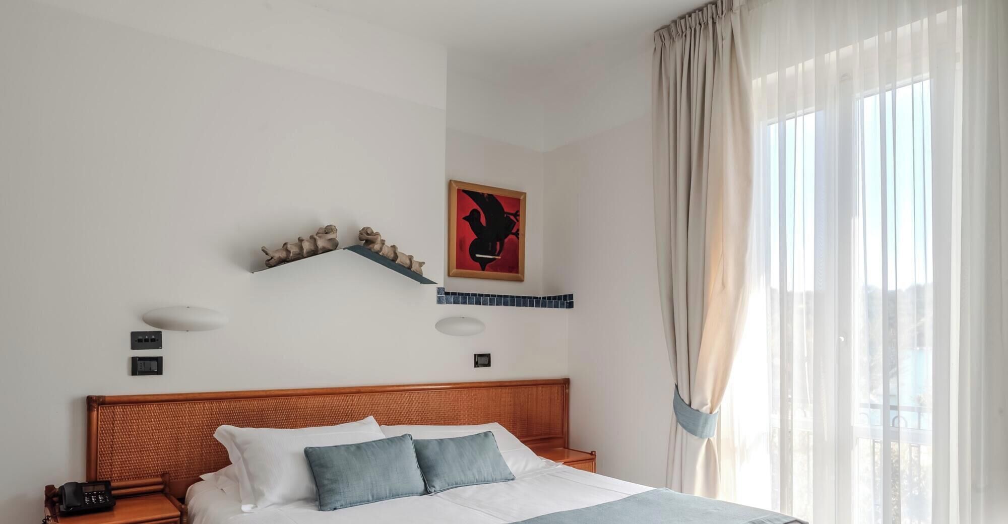 A hotel room with a bed, a telephone, a wall-mounted shelf with decorative items, a painting, and a window with curtains.