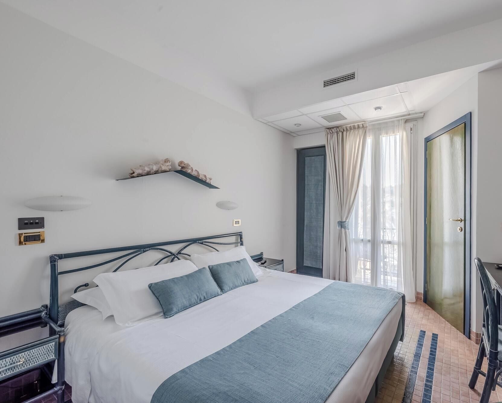 Een hotelkamer met een bed, een nachtkastje, een wandplank, een raam met gordijnen en een deur.