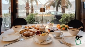 Mesa de café da manhã com pães, frutas e bebidas, vista para palmeiras e um carro do lado de fora.