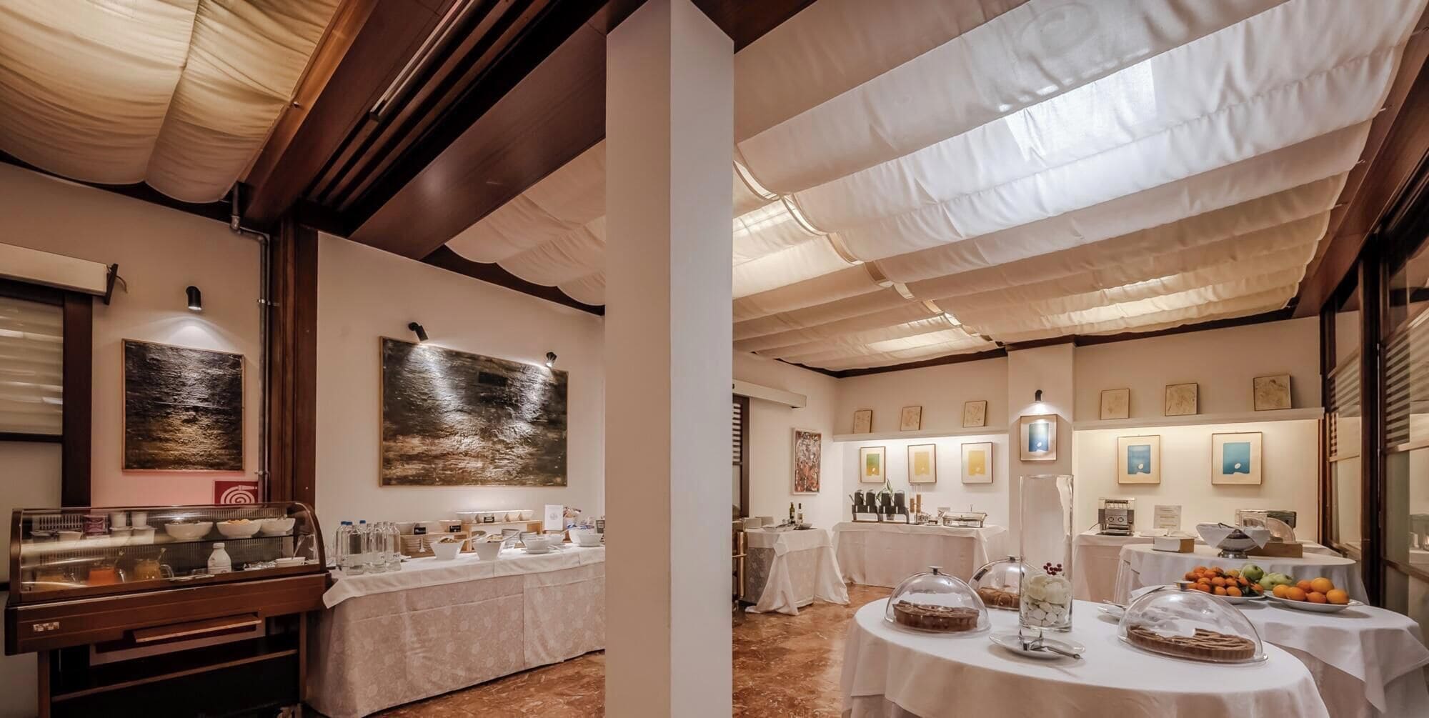 Comedor de hotel con mesas dispuestas para el desayuno, obras de arte en las paredes y una barra de buffet.