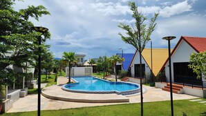 A swimming pool surrounded by a well-maintained lawn, with modern buildings featuring colorful roofs in the background. - LML Kampot Relax House (Kampot)