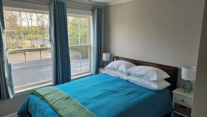 Un dormitorio con una cama, un ventilador de techo, una ventana con cortinas y una mesita de noche con lámpara.