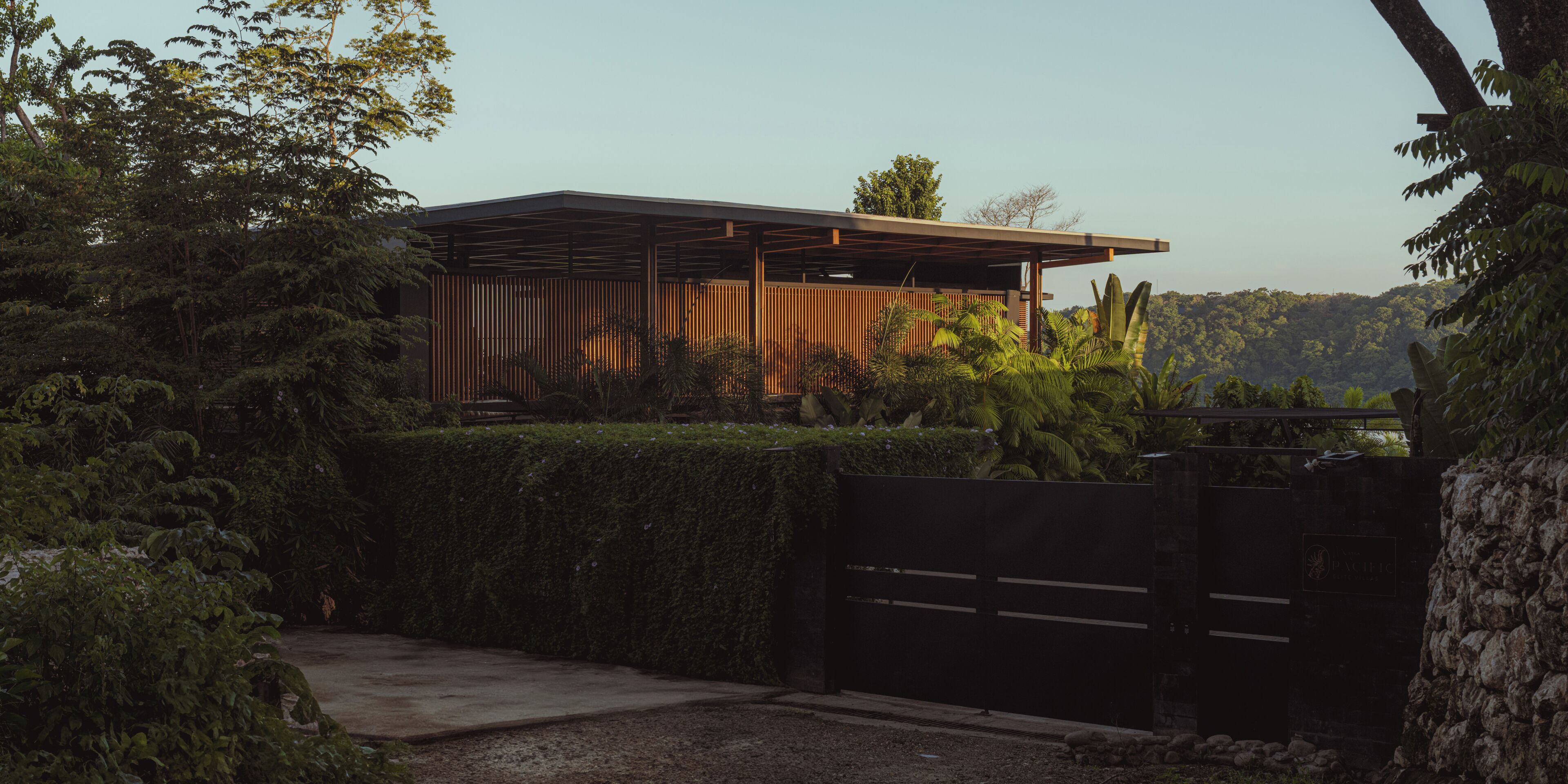 A modern building with a flat roof, surrounded by dense greenery and a gated entrance.