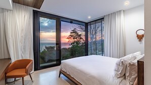 Ein Schlafzimmer mit einem großen Bett, einem Sessel und Blick auf den Sonnenuntergang durch eine Glastür.