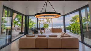 Ein modernes Wohnzimmer mit einem großen beigefarbenen Sofa, einem Essbereich mit Stühlen und Meerblick durch große Fenster.