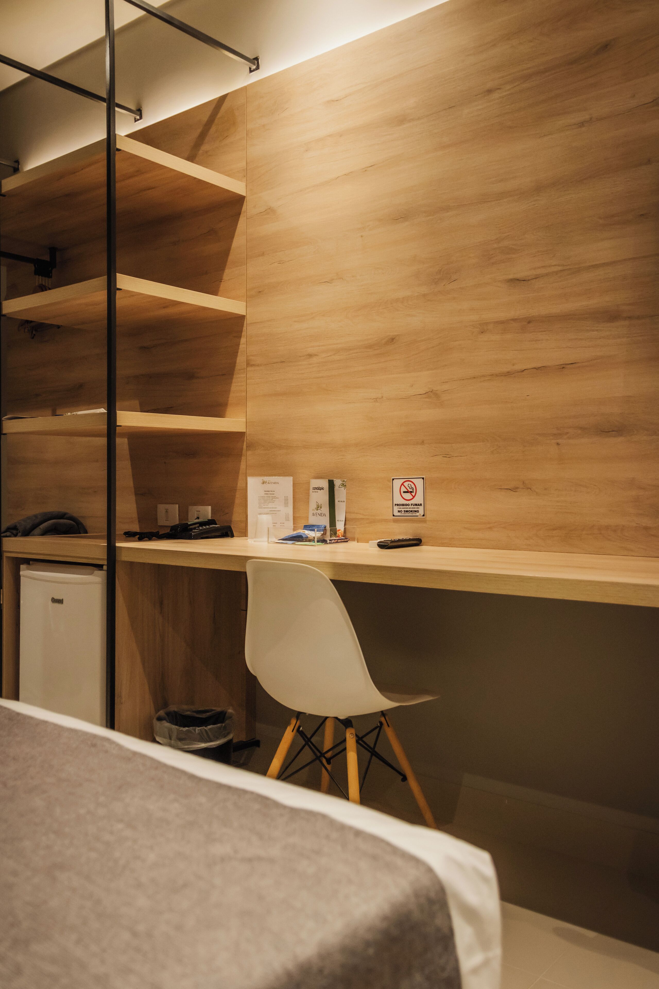 A compact hotel room with a wooden desk, a white chair, a small refrigerator, and a bed with a grey blanket.