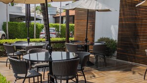 An outdoor patio with round tables and chairs, surrounded by palm trees and a wooden deck. - Hotel Avenida (Sorriso)