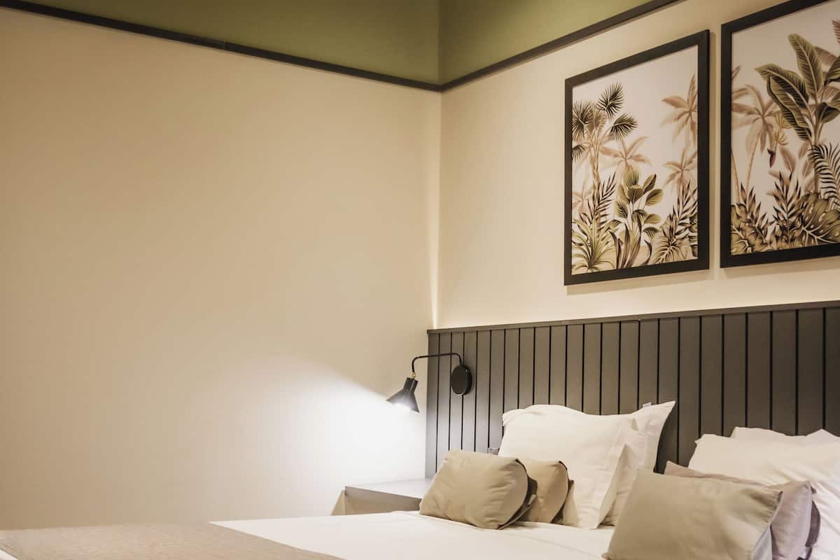 Una cama bien tendida con una colcha acolchada, una lámpara de noche y dos láminas enmarcadas con motivos botánicos en la pared.