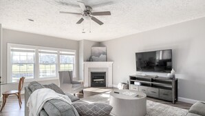 A spacious living room with a fireplace, a large flat-screen TV, and a ceiling fan. - Relax in Style, Golf course Community Townhouse - 3440 (Baldwinsville)