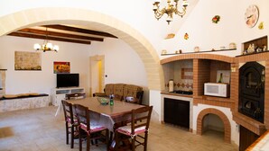 A dining area with a table and chairs, a television, and a kitchen with a brick oven. - Trulli del Capo - Casa Tipica (Monopoli)