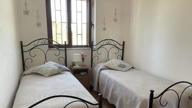 Ein Schlafzimmer mit zwei Einzelbetten, einem Nachttisch mit Lampe und Blumen sowie einem Fenster mit Gitterstäben.