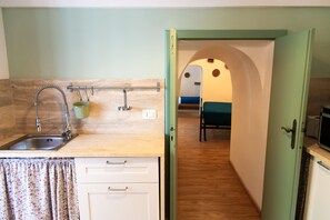 A compact kitchen with a sink, faucet, and a hanging pot rack. A door leads to a bedroom with a bed and a small table. - Trulli del Capo - Trulli (Monopoli)