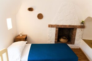 A bedroom with a bed, a small wooden nightstand, a white wall, a stone fireplace, and a window. - Trulli del Capo - Trulli (Monopoli)