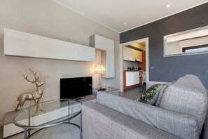A modern living room with a sofa, a glass coffee table, a flat-screen TV, a decorative deer sculpture, and a kitchen visible through an open doorway.