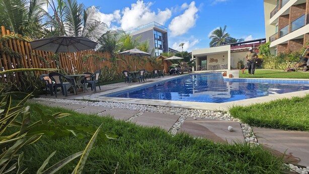 Una piscina rodeada de mobiliario exterior y vegetación, con un edificio moderno al fondo.