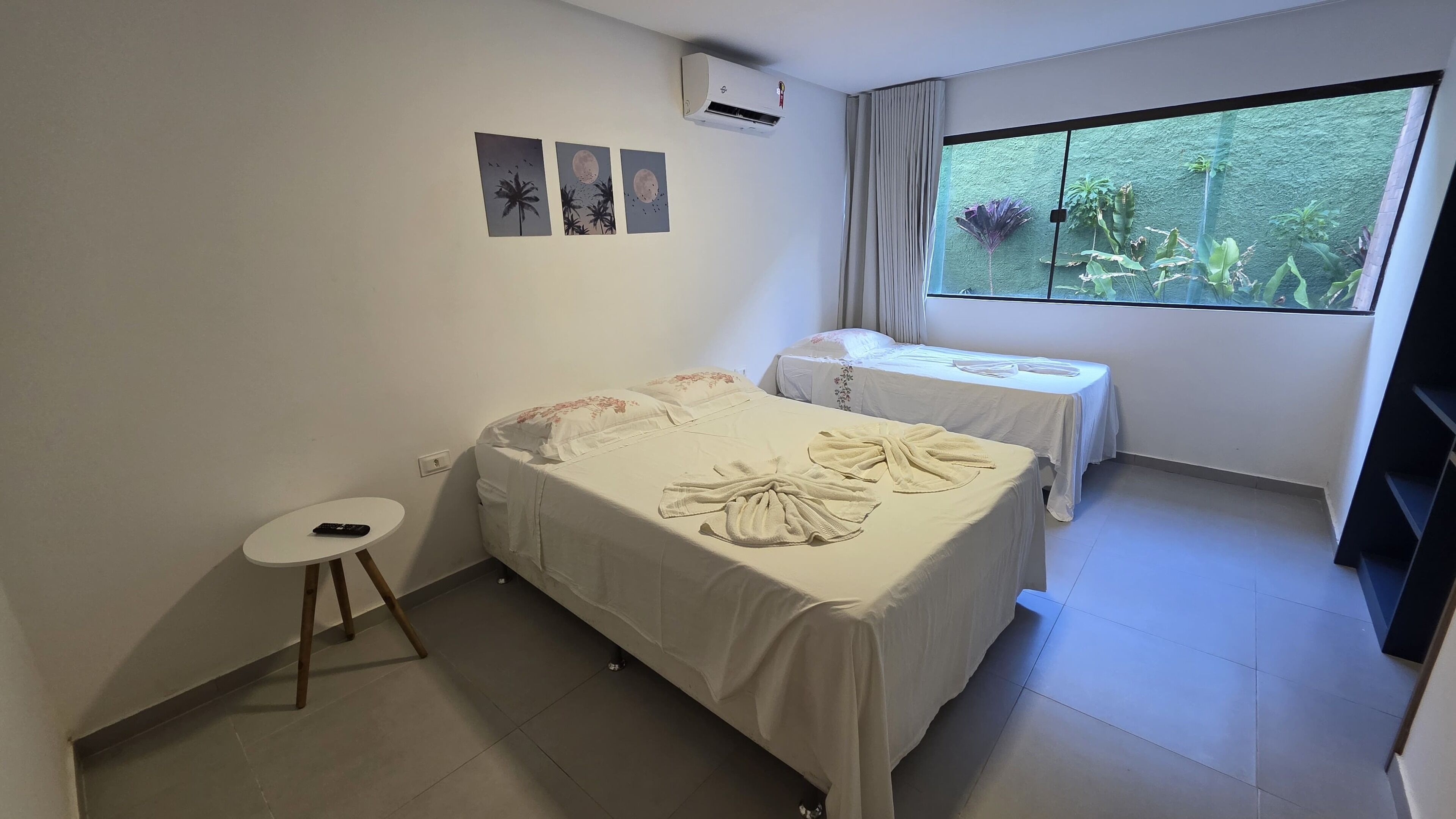 A hotel room with two beds, a small table, and a window with a view of greenery.