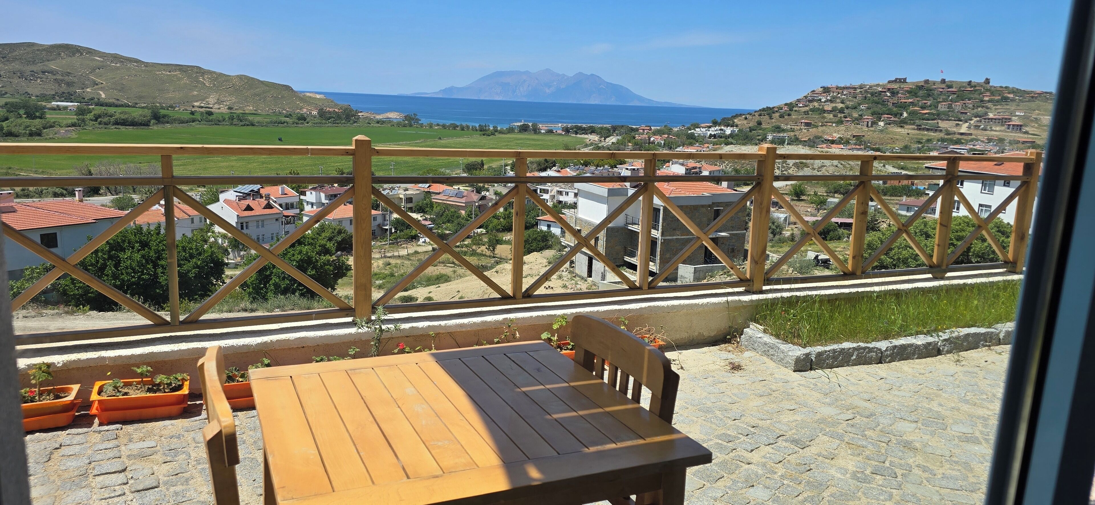 A balcony with a wooden table and chairs, overlooking a coastal town with buildings and a mountain in the distance.