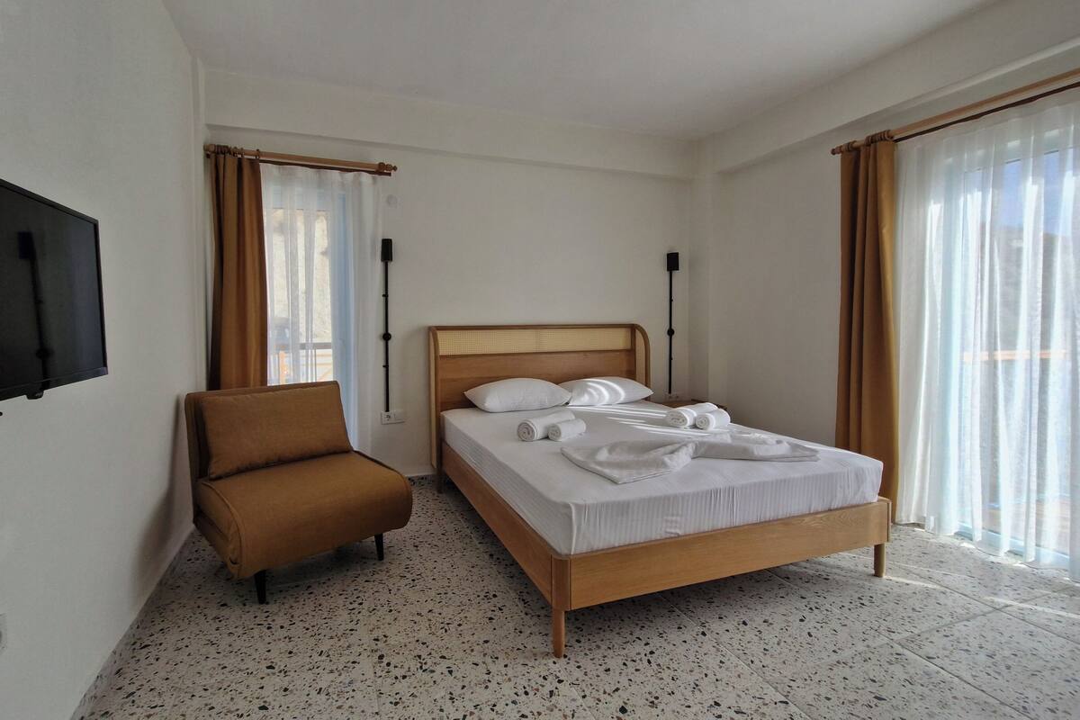 A hotel room with a bed, a chair, a TV, and large windows with curtains.