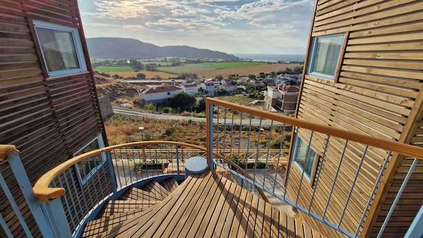Une terrasse en bois avec un escalier en colimaçon menant à un balcon d’où l’on peut admirer un paysage rural avec des maisons et des champs.