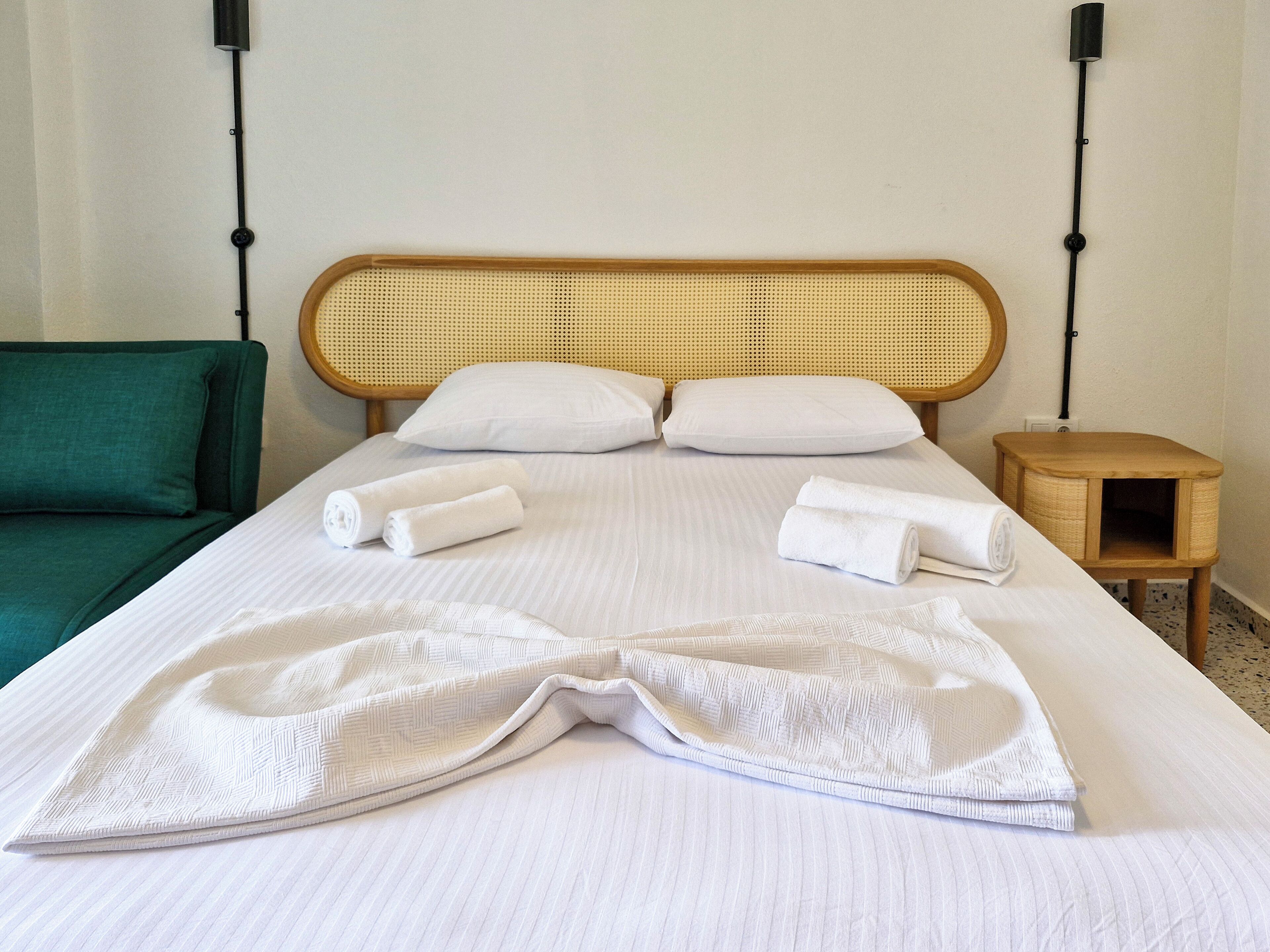 Un lit bien fait, avec du linge de lit blanc et des serviettes pliées, un canapé vert et une petite table de chevet en bois.