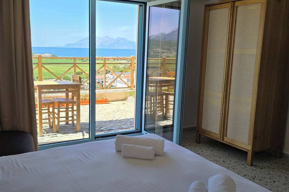 A hotel room with a bed, a TV, a wardrobe, and a balcony with a view of the sea and mountains.