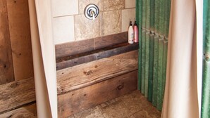 Un baño de cabaña de madera con zona de ducha con azulejos de piedra, un banco de madera y una cortina verde.