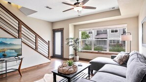 A spacious living room with a gray sofa, a wooden coffee table, a TV on a stand, a ceiling fan, and a large window with a view of a house. - Old Town North Getaway – King Beds & Game Room! (Fort Collins)