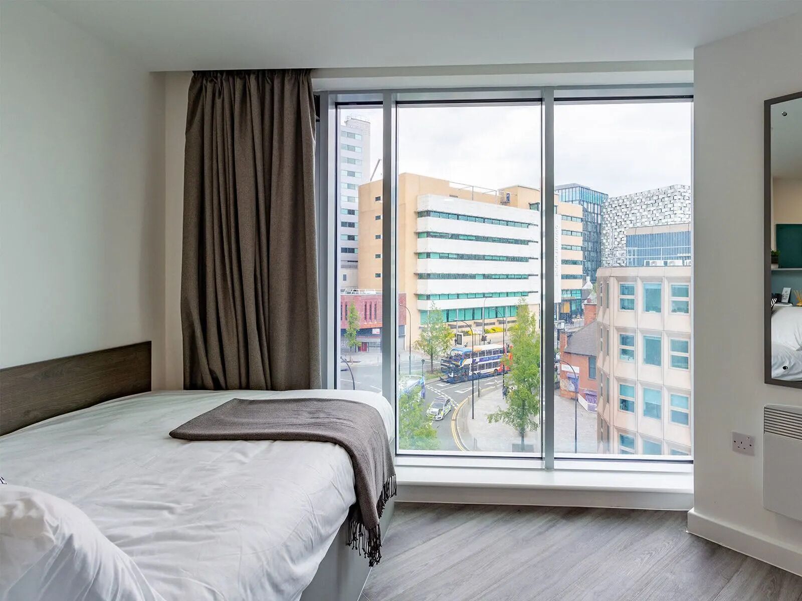 Ein Hotelzimmer mit großem Fenster, einem Bett, einem Nachttisch und Blick auf die Stadt.