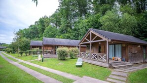 A row of rustic cabins with wooden exteriors and pitched roofs, surrounded by lush greenery and a paved walkway. - ZEcamping 4 étoiles piscine chauffée chalet TV LV BBQ 35m2 (Aramits)