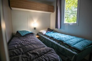 Una habitación pequeña y funcional con dos camas individuales, una mesita de noche, una lámpara en la pared y un ventanal con cortinas.