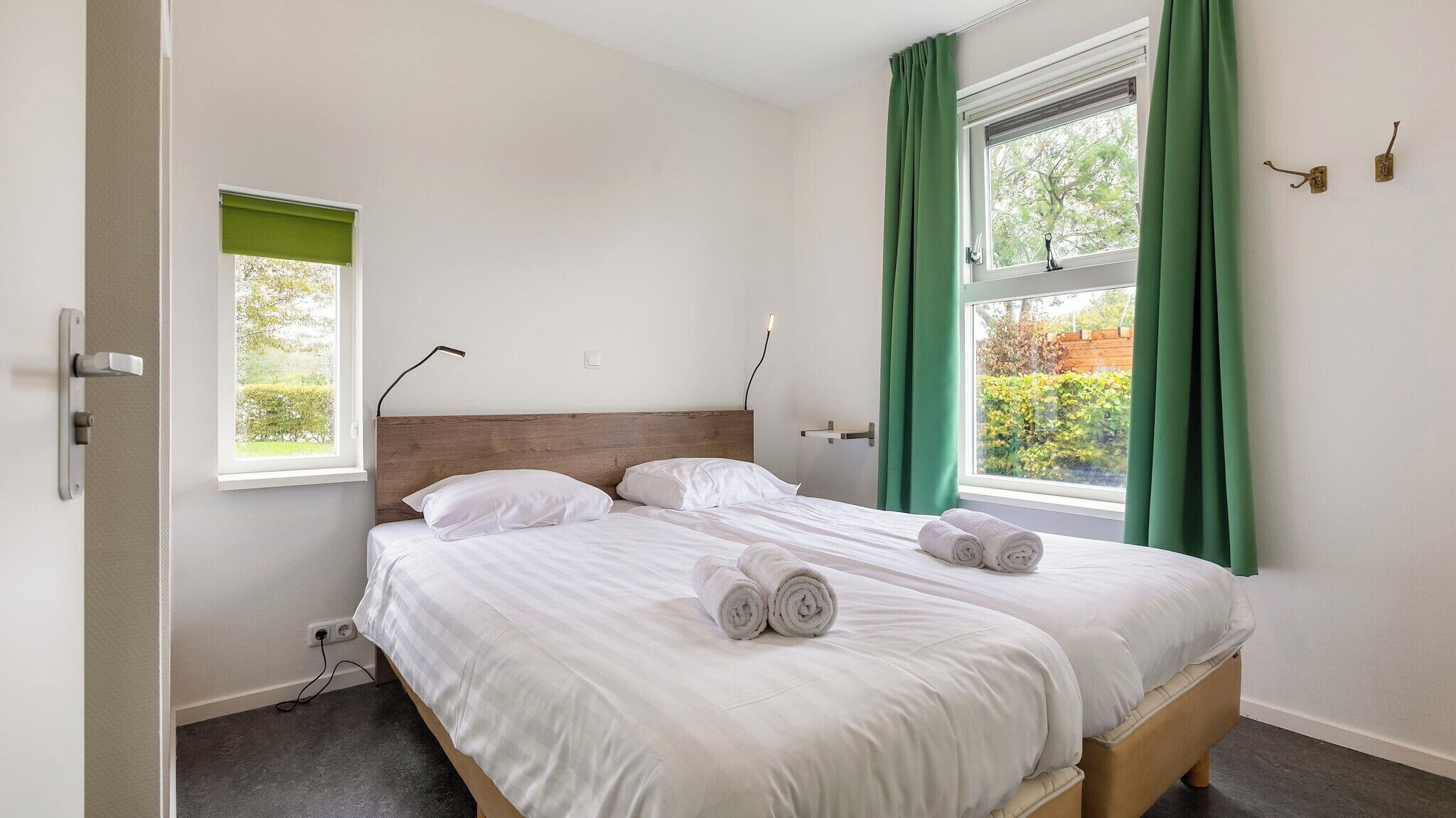 A bedroom with a wooden headboard, two white beds, green curtains, and a view of greenery outside.