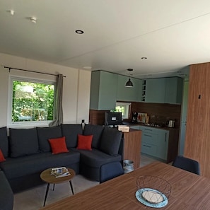 Un espace de vie moderne comprenant un canapé, une table basse et une table à manger. On aperçoit à l’arrière-plan un coin cuisine avec des placards et des appareils électroménagers.
