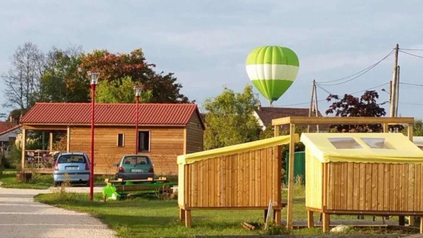 Eine Holzhütte mit rotem Dach, im Hintergrund ein grün-weißer Heißluftballon und eine gelbe Konstruktion mit einem Fenster.
