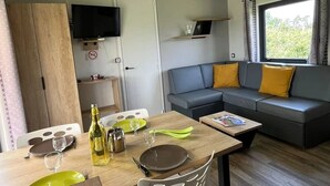 Una sala de estar moderna con comedor, sofá gris con cojines amarillos, un televisor de pantalla plana y una ventana con cortinas.