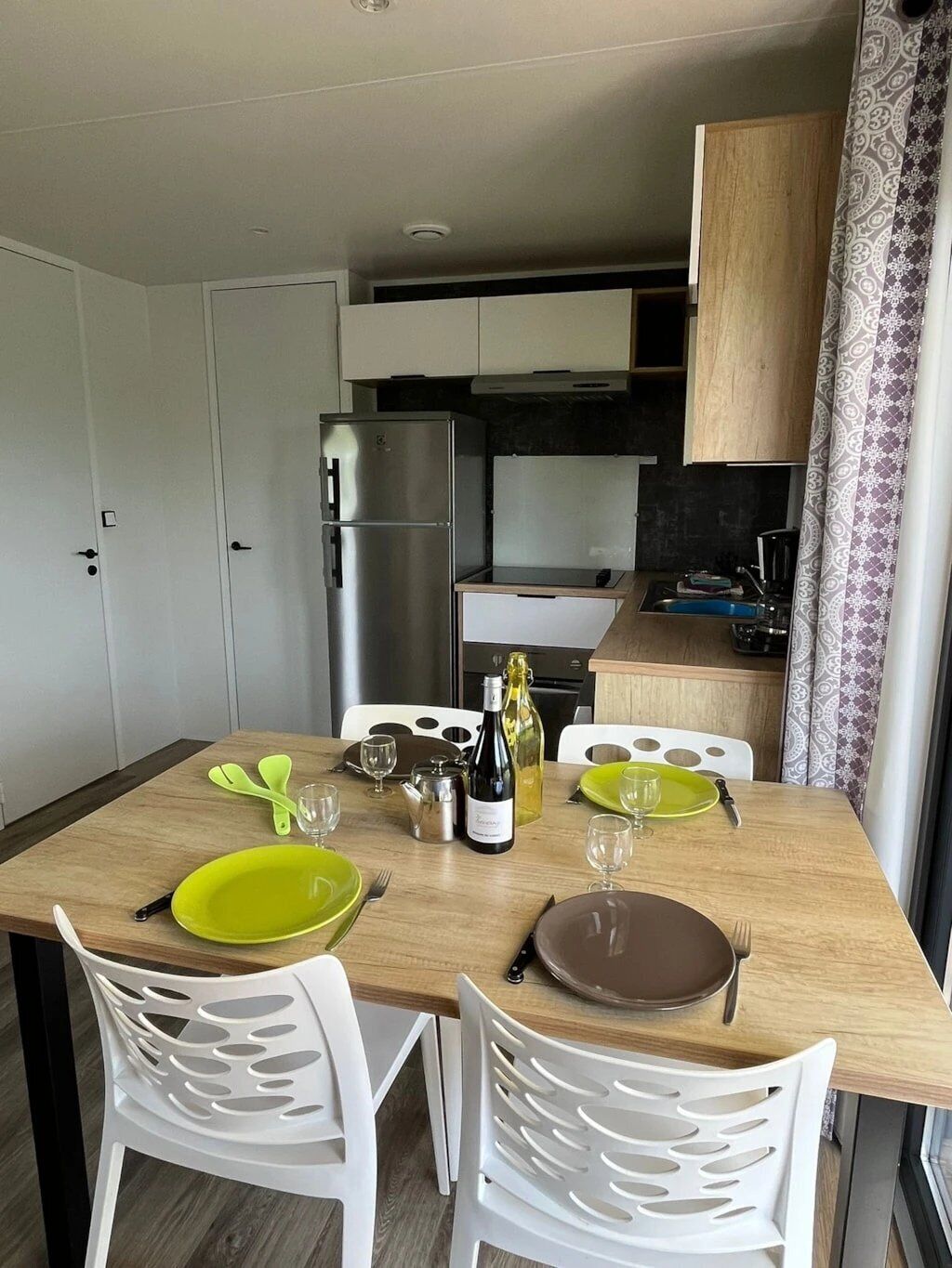 Cocina compacta con armarios blancos, un refrigerador de acero inoxidable y una mesa de comedor con platos, vasos y una botella de vino.