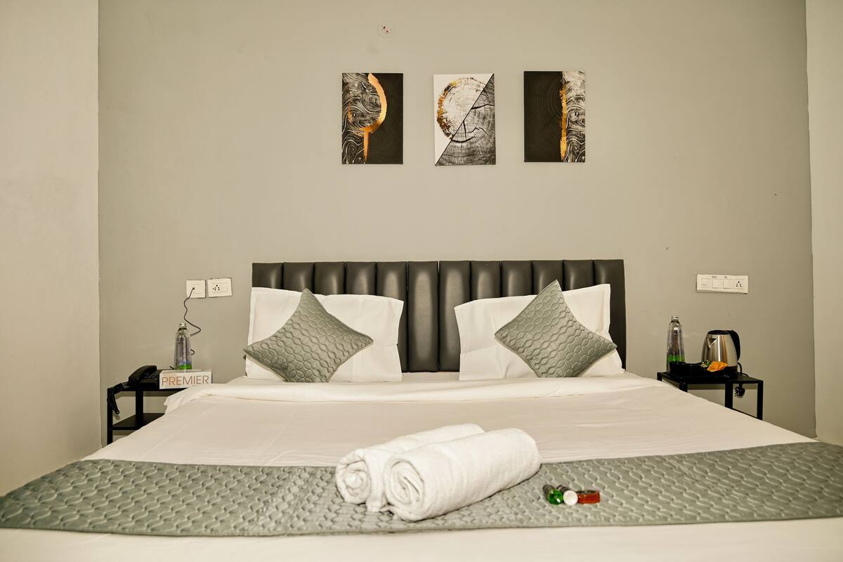 A neatly made bed with a grey quilt, two pillows, and a rolled white towel on a grey bedspread. There are three wall-mounted artworks above the bed.