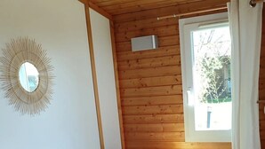 A cozy room with a wooden ceiling, a bed with white and green pillows, a small table with a bottle and glasses, and a window with white curtains.