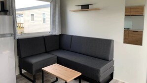 A compact living space with a gray sofa, a small wooden table, a window with curtains, and a kitchen area visible through an open door. - ZEcamping 3 étoiles familial piscine chauffée locatif TV BBQ 32m2 (Grand-Laviers)