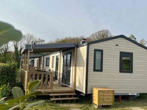 A single-story mobile home with a porch and a small deck, surrounded by greenery.