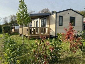 A mobile home with a deck, surrounded by greenery and a clear sky.